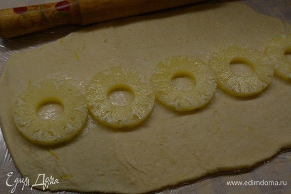 Стол застелить пищевой пленкой (так с тестом лучше работать). Достать тесто и раскатать его в прямоугольный пласт. На середину уложить кружочки ананасов.