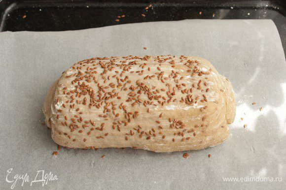 Накройте хлеб, чтобы уберечь от заветривания, и отправьте в теплое место на 50-60 минут. За это время буханка вырастет. Переверните хлеб на противень аккуратно, чтобы воздух не вышел. Сбрызните буханку водой и посыпьте льном.