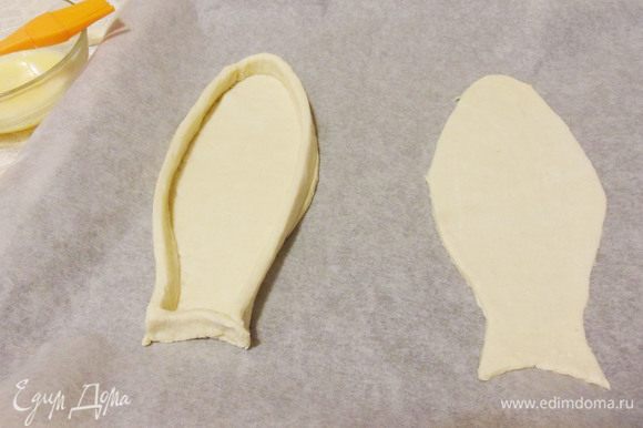 Переложить заготовки на противень застеленный бумагой для выпечки. Из обрезков теста вырезать длинные полоски, смазать их с одной стороны яйцом. Выложить полоски «бортиками» по краю рыбки, смазанной стороной вниз.