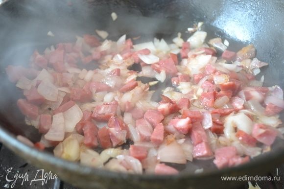Как только лук станет румяниться, добавьте мелко нарезанную копченую колбаску. Жарьте минуту, помешивая. Колбаса отдаст свой аромат копчения. Затем добавьте измельченный чеснок.