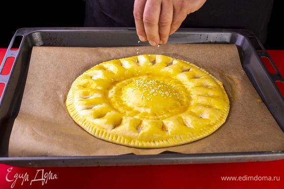 Затем надрежьте тесто, формируя лучики солнца. С помощью круглой формы небольшого диаметра обозначьте центр. Дайте пирогу отдохнуть и насстояться 10 минут. Взбейте яичный желток и смажьте им пирог, используя силиконовую кисточку. Верх пирога посыпьте семенами кунжута.