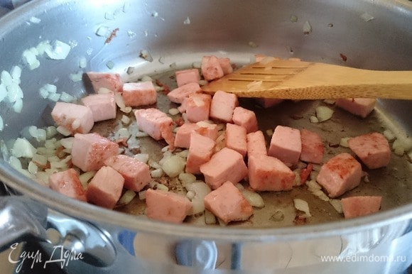 Нагрейте масло в большой сковороде. Положите нарезанную колбасу и жарьте на среднем огне. К чуть обжаренной колбасе добавьте лук и готовьте до золотистого цвета около 3 минут.