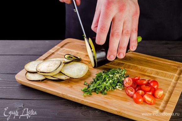 Баклажаны нарежьте тонкими кружочками, помидоры черри разрежьте пополам. Измельчите чеснок и зелень.