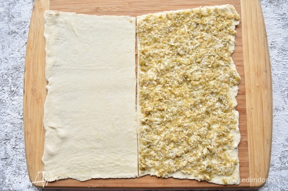Слоеное тесто раскатать в прямоугольник и разрезать его на две равные части. На одну из них выложить подготовленную начинку и распределить по всей поверхности.