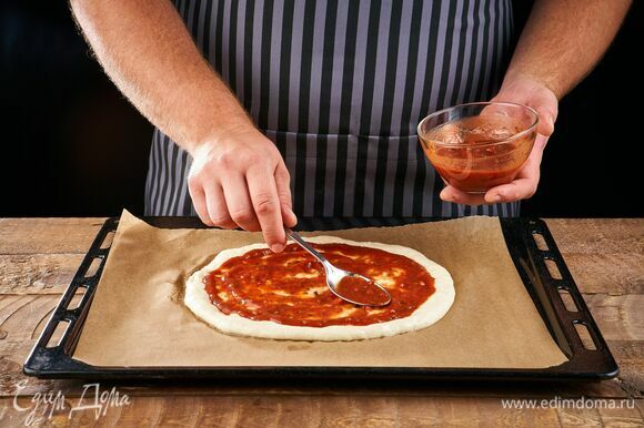 Раскатайте тесто в круглую форму, сформировав небольшие бортики, выложите на противень, застеленный пергаментом. Смажьте основу томатным соусом.