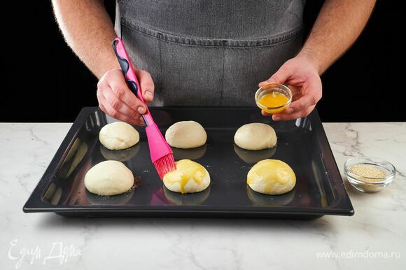 Из поднявшегося теста сформируйте шесть булочек. Выложите заготовки на противень, смажьте каждую яичным желтком и посыпьте кунжутом. Запекайте в духовке, разогретой до 190°C, около 18 минут.